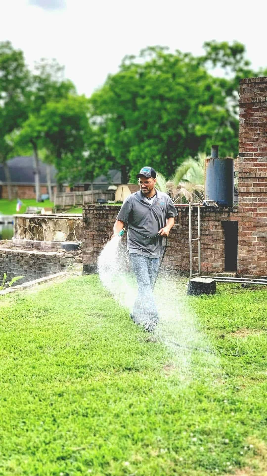 A man wearing a cap and glasses waters a green lawn with a garden hose in a backyard, showing how reliable lawn care in your area can keep yards lush near brick structures and trees.