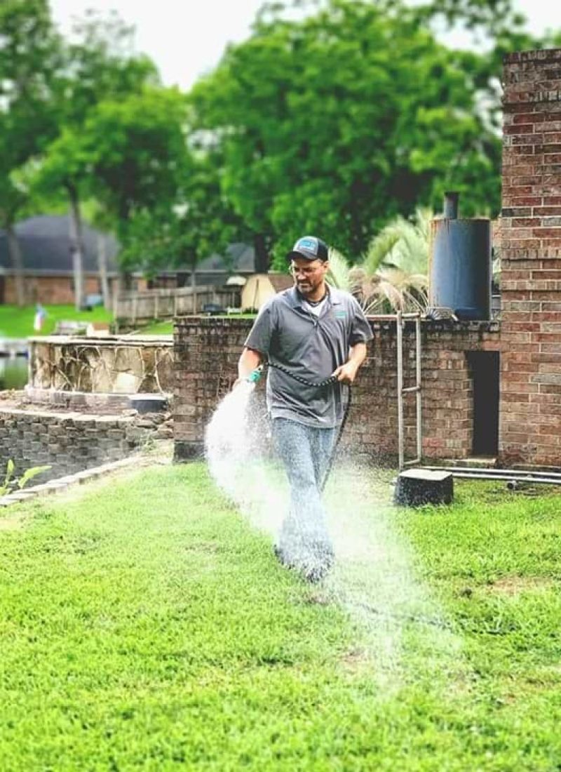 A man wearing a hat and casual clothing is watering a lawn using a hose. In the background, a brick structure stands tall with trees and a lake visible, making the scene look like something right out of Scooby-Doo's many mysteries.